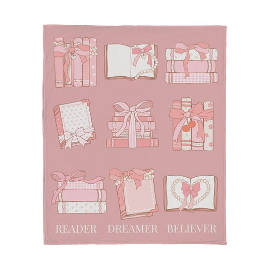 Pink fleece blanket with coquette-style grid of books and bows, and the words 'Reader, Dreamer, Believer' at the bottom.